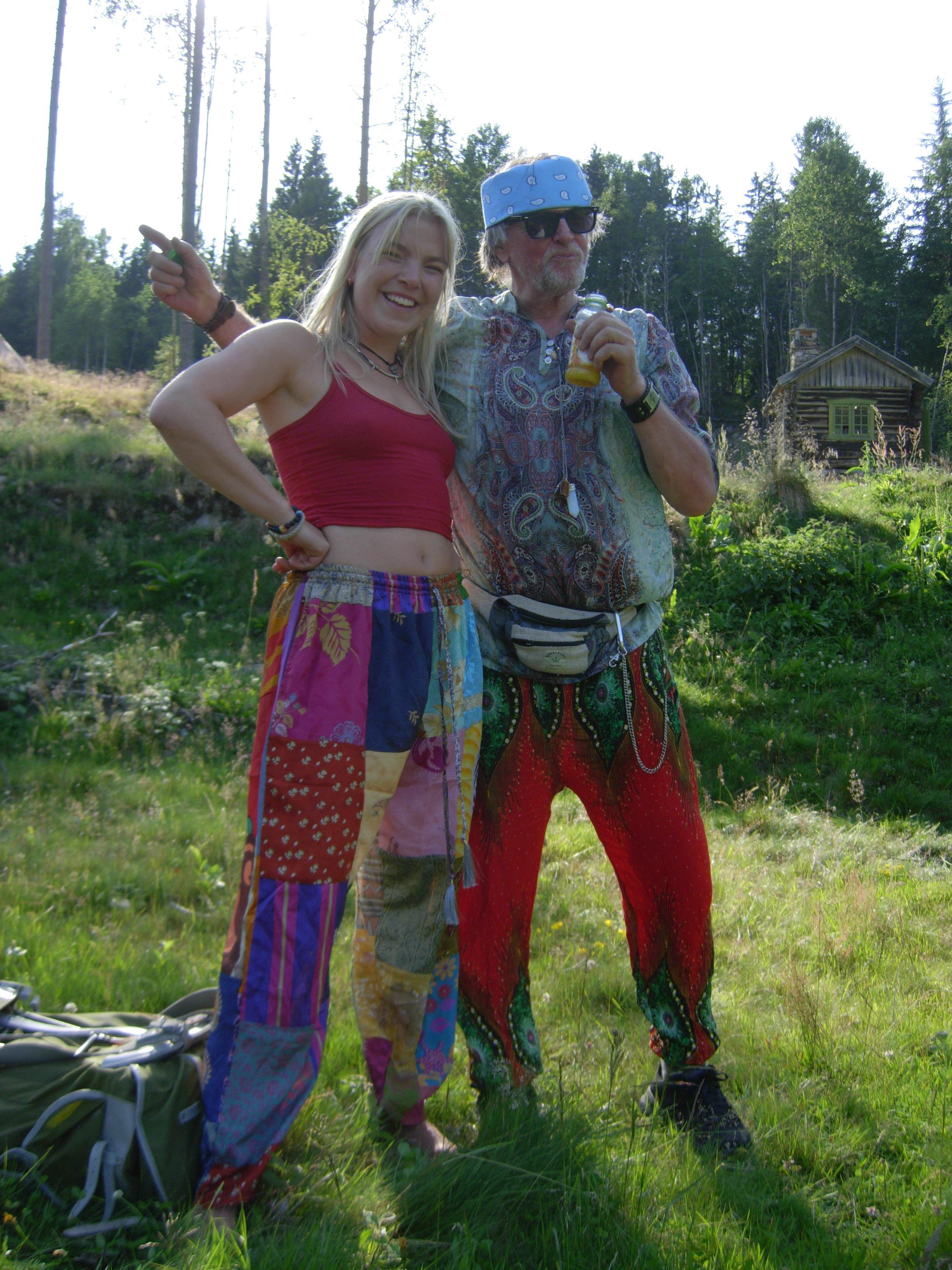 To festivaldeltagere i fargerike klær på sommereng med tømmerhytte og furuskog i bakgrunnen