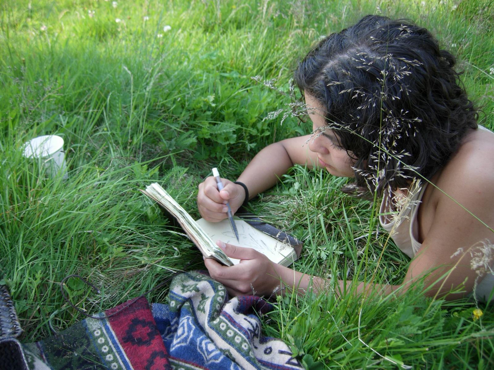 Person som ligger i gresset og skriver i dagbok, omgitt av grønt gress og et fargerikt teppe